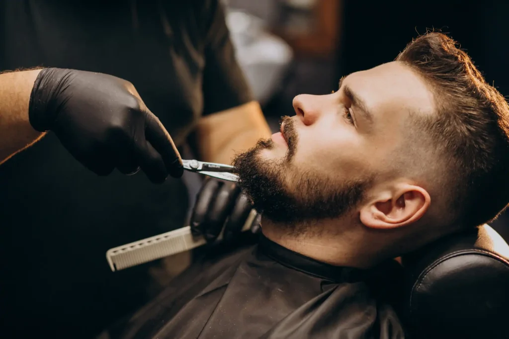 Barber w czarnych rękawiczkach przycina brodę klienta.