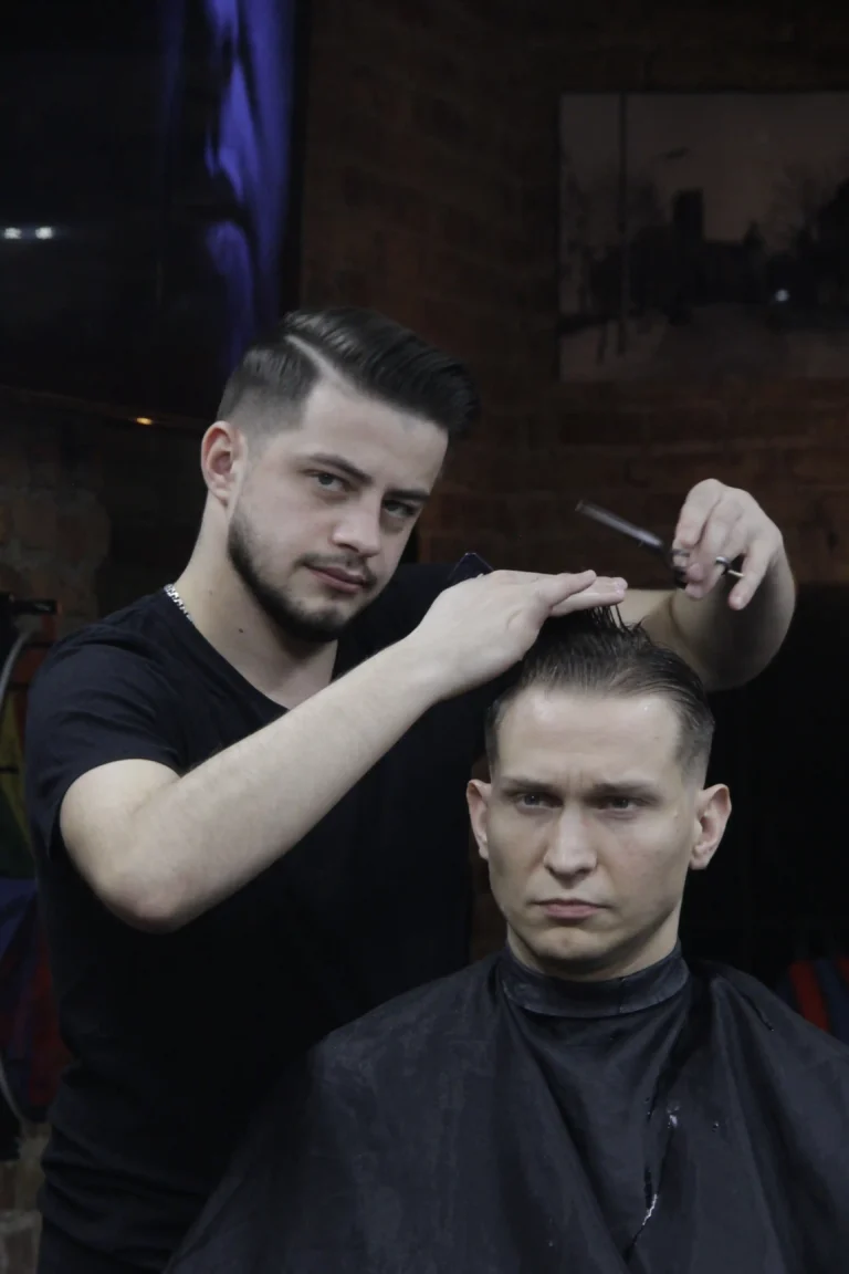 Fryzjer strzyże włosy klientowi nożyczkami, patrząc w kamerę.