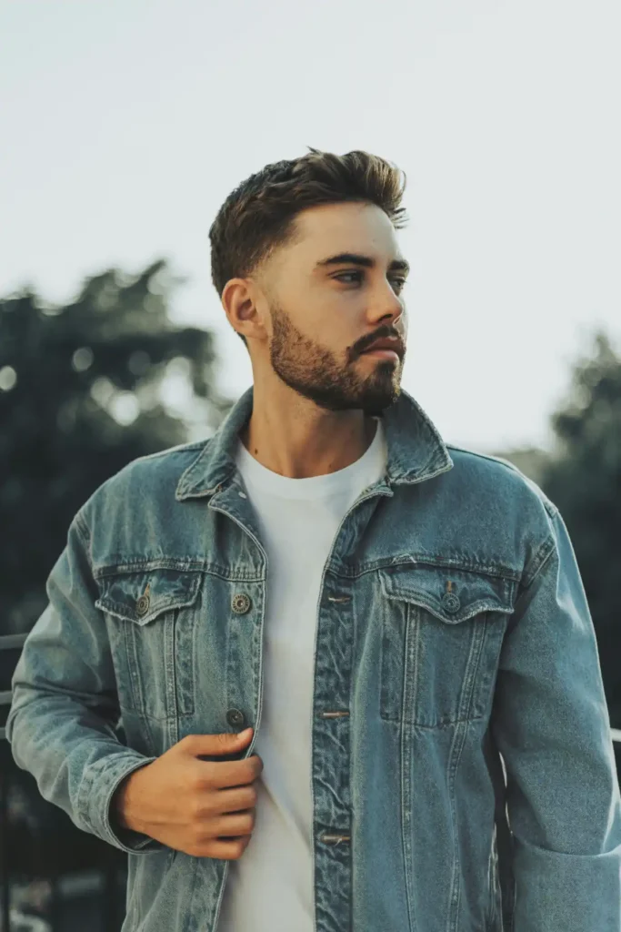 Mężczyzna w białym T-shircie i jeansowej katanie pozuje na zewnątrz.